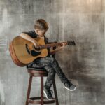 A Young Boy Sitting on a Wooden Chair while Playing Guitar