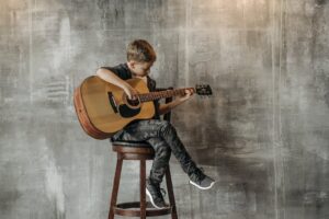 A Young Boy Sitting on a Wooden Chair while Playing Guitar